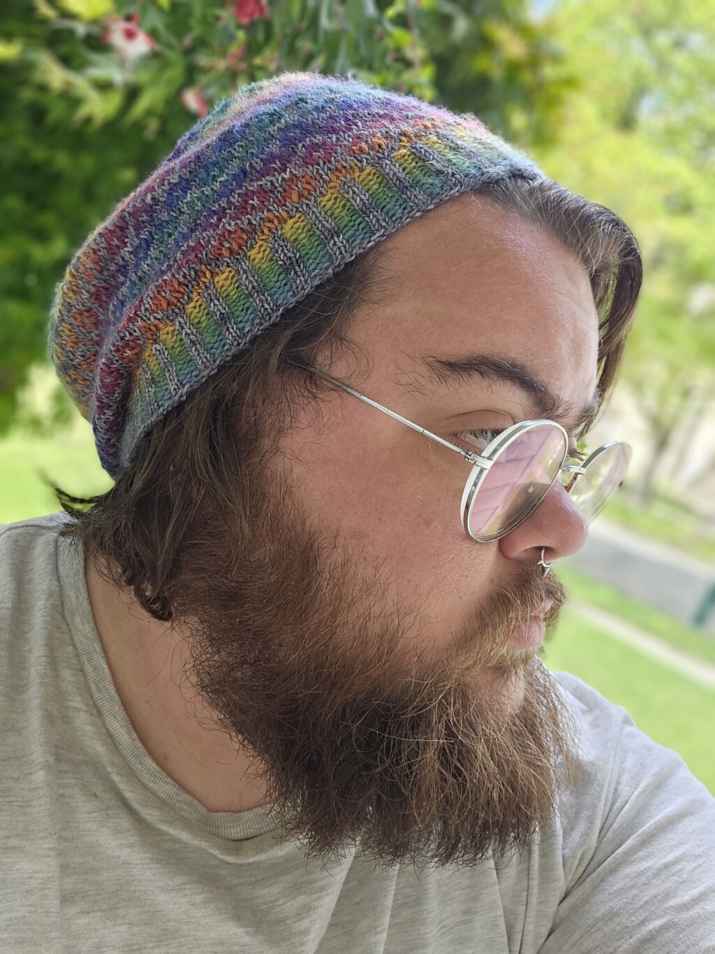 Image description: A bearded man wearing glasses and a hand knit hat. The hat is a design of interlocking diamonds in a rainbow gradient and a silver for contrast.The background is full of green plant life. End ID
